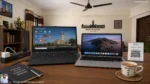 Laptop and MacBook on wooden desk in Bhavnagar home with ceiling fan above, showing desktop with essential software icons, UPI scanner and chai cup beside, power strip with surge protector