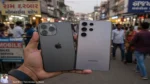 A hand holding an iPhone and a Samsung Galaxy phone side by side in a busy Indian market street